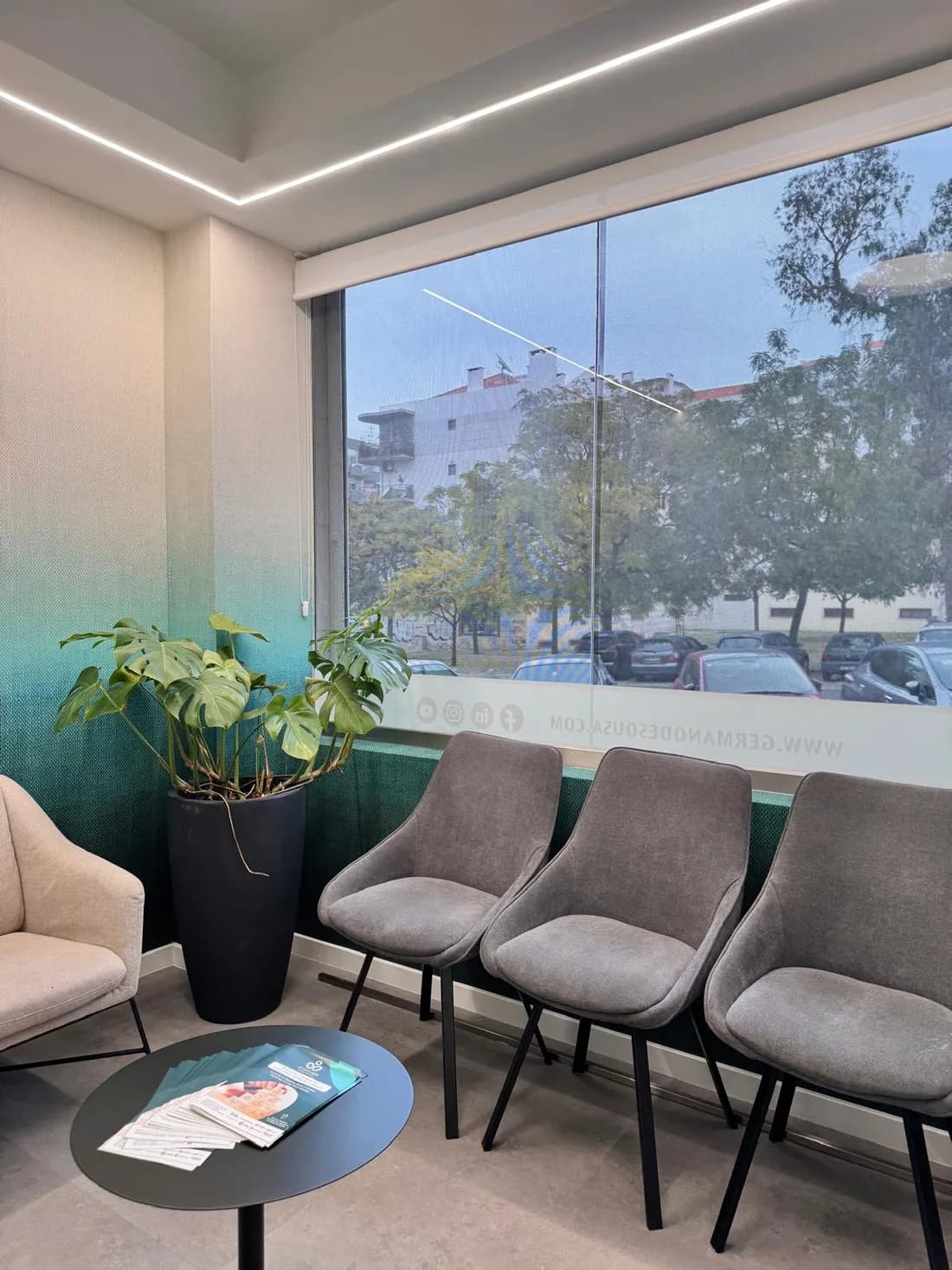 Sala de espera com cadeiras junto à janela e plantas, banhada em luz natural.